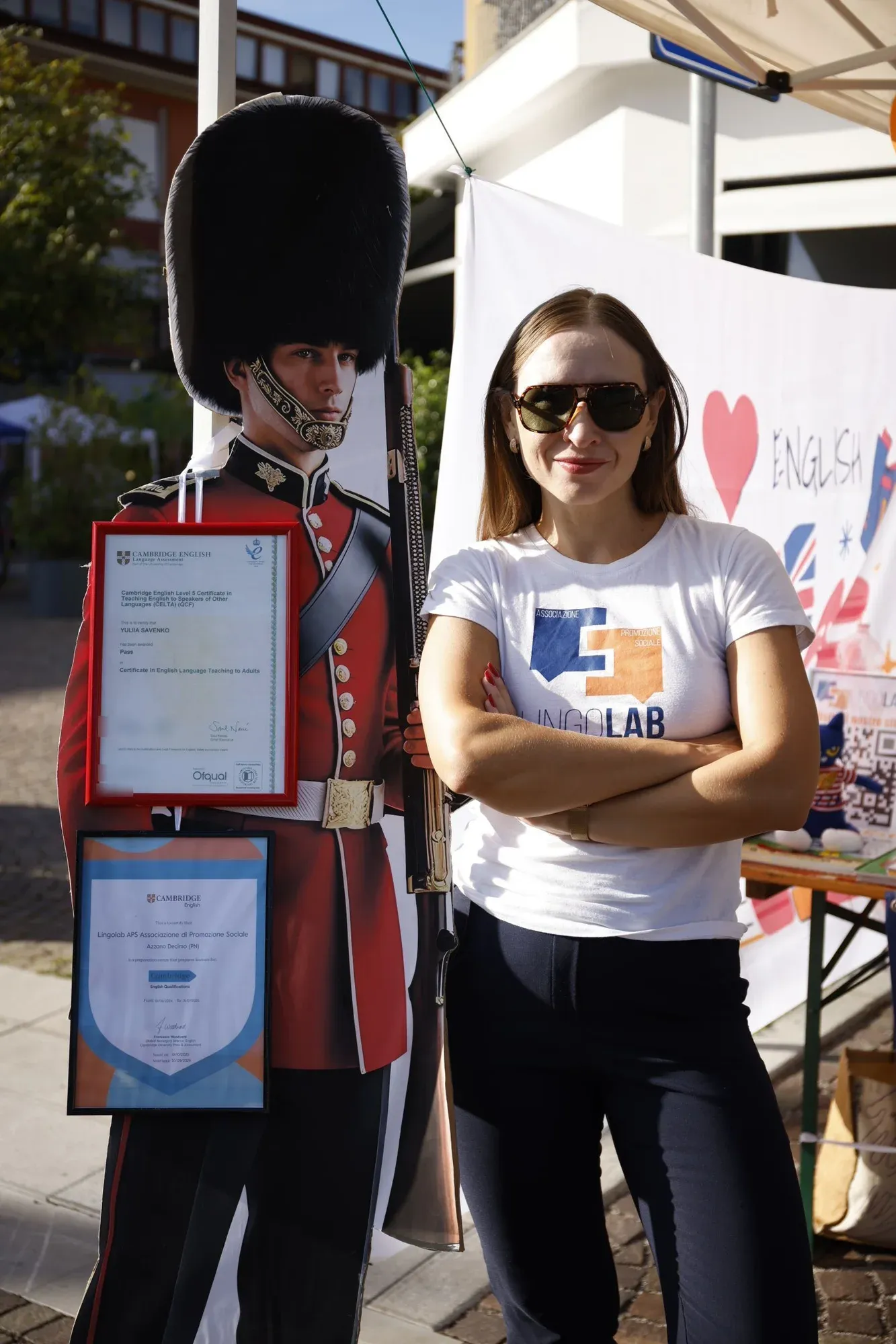 The teacher at the stand next to a cut-out of a British Royal Guard holding Cambridge certificates