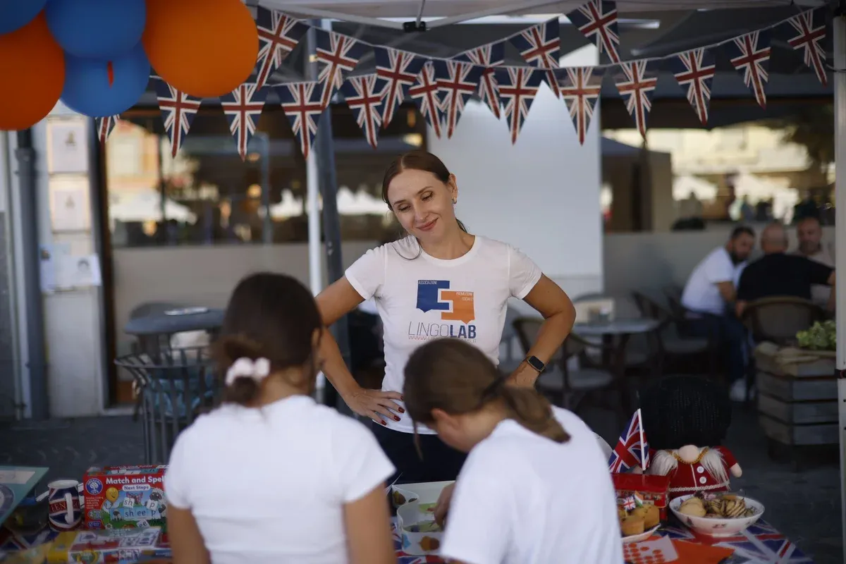 The teacher at the LingoLab stand at Azzano in Festa, under Union Jack bunting, with two girls at the table